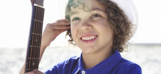 Un jeune enfant souriant avec des boucles et un chapeau tenant une guitare, évoquant le bonheur lié à l'épanouissement auditif.