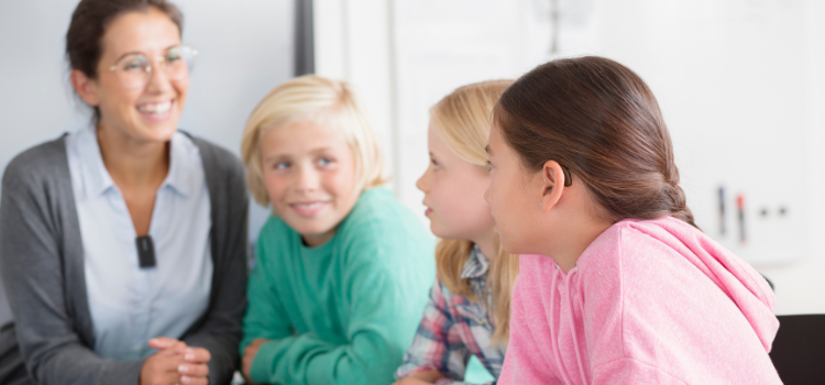 Trois enfants assis en cours à côté de leur professeur qui leur sourit.