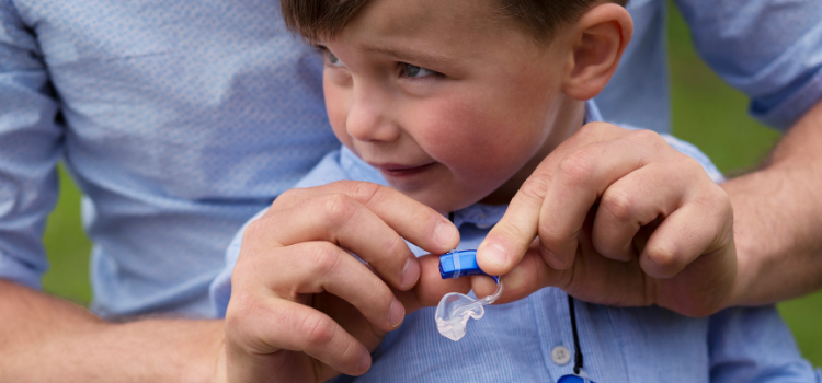 Un enfant souriant reçoit de l'aide pour ajuster son appareil auditif bleu.