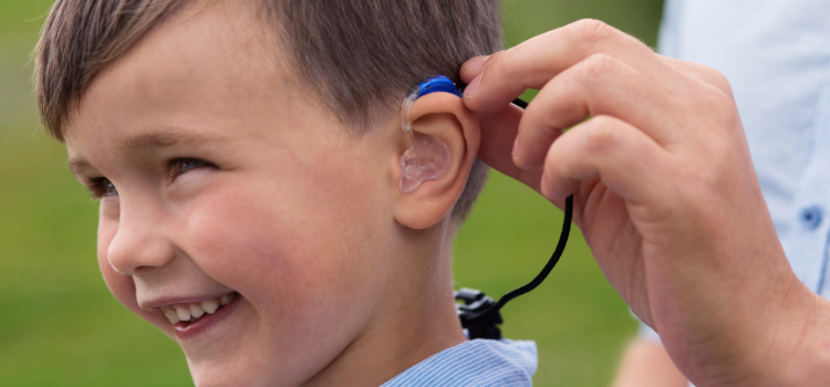 Un enfant souriant reçoit de l'aide pour ajuster son appareil auditif bleu.